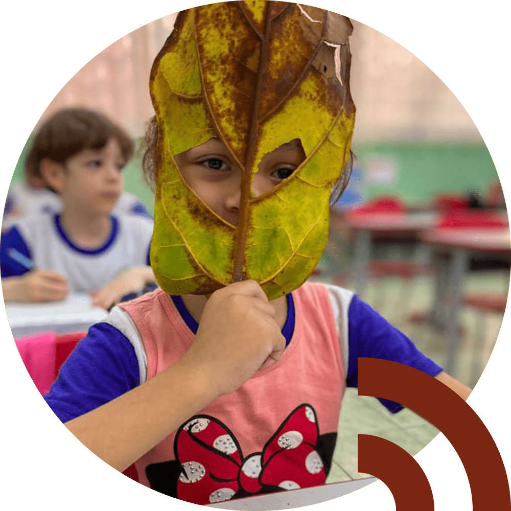 2. Menina com uma folha sobre o rosto, sentada em uma sala de aula, com colegas ao fundo, em atividade do projeto Caminhos para a Cidadania (agora Escolas baseadas na Natureza), realizado em parceria técnica com o Crescer.